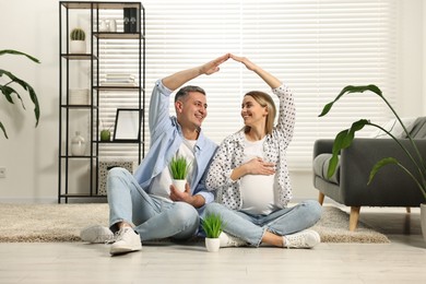 Young family housing concept. Pregnant woman with her husband forming roof with their hands while sitting on floor at home