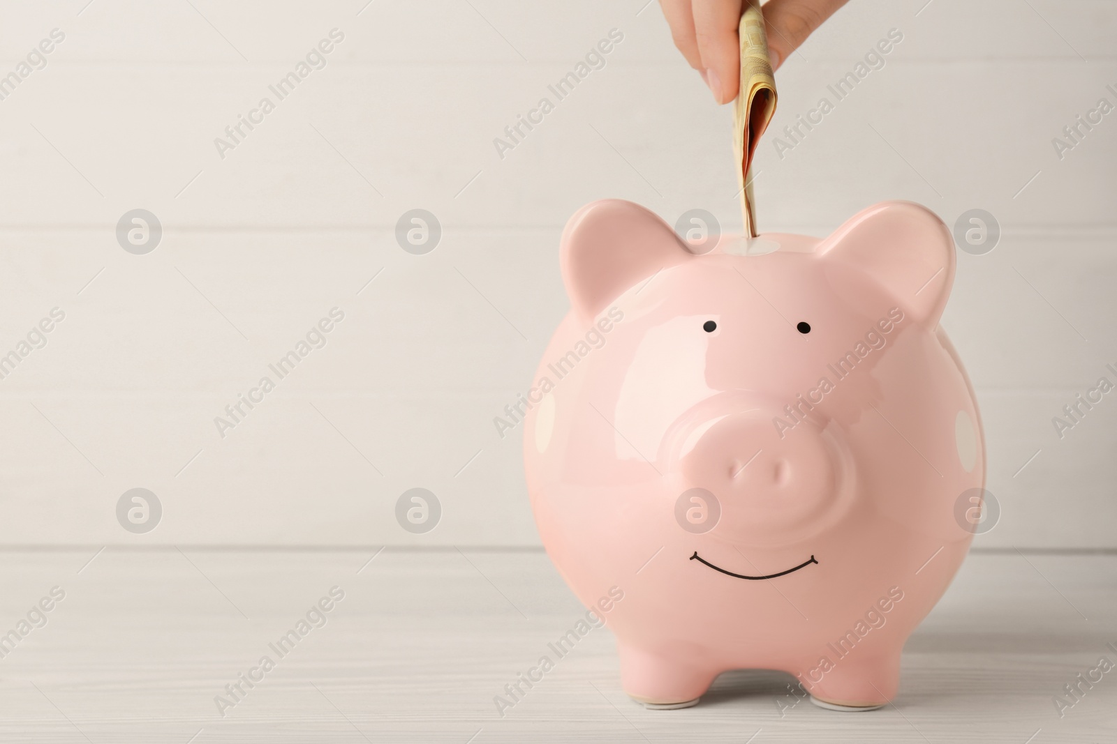 Photo of Woman putting banknotes into piggy bank at white wooden table, closeup. Space for text