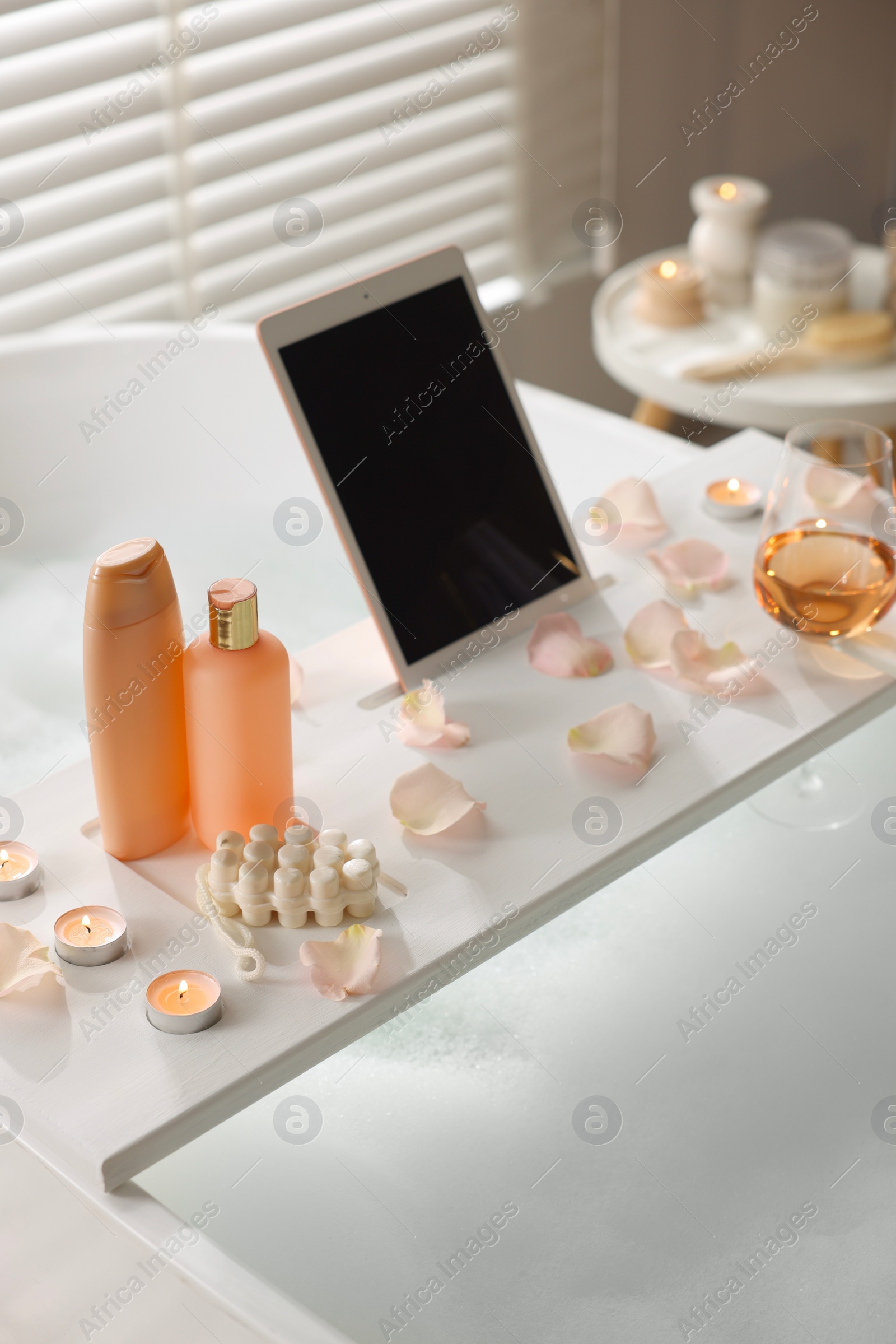 Photo of Wooden tray with tablet, wine, toiletries and flower petals on bathtub in bathroom