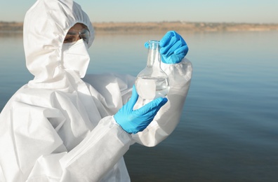 Scientist in chemical protective suit with florence flask taking sample from river for analysis