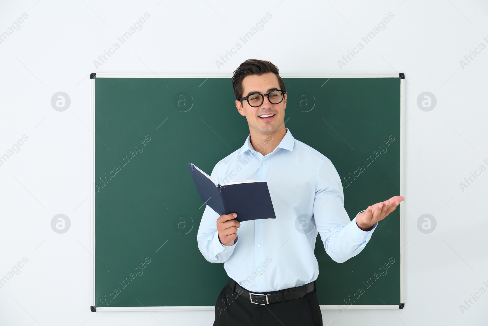 Photo of Young teacher with book near blank chalkboard in classroom