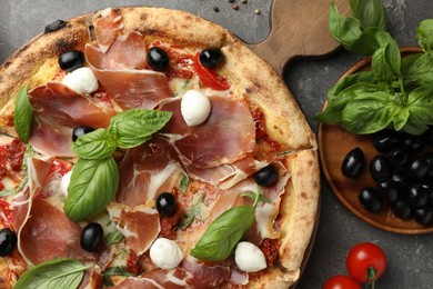 Photo of Tasty pizza with cured ham, olives, mozzarella cheese, tomatoes and arugula on gray table, flat lay