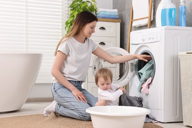 Photo of Happy mother with her daughter putting baby clothes into washing machine in bathroom