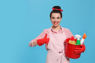 Young housewife holding bucket with cleaning supplies on light blue background, space for text
