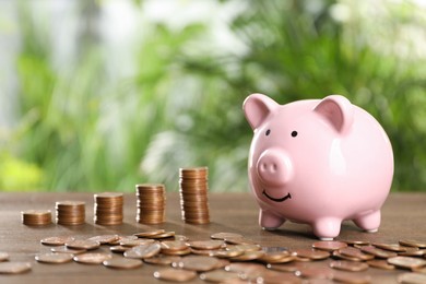 Many metal coins and piggy bank on wooden table against blurred green background. Space for text