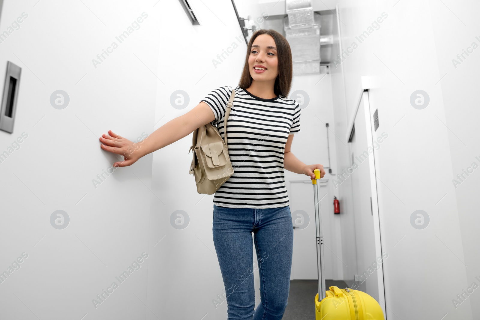 Photo of Happy young woman with backpack and suitcase walking along hostel corridor