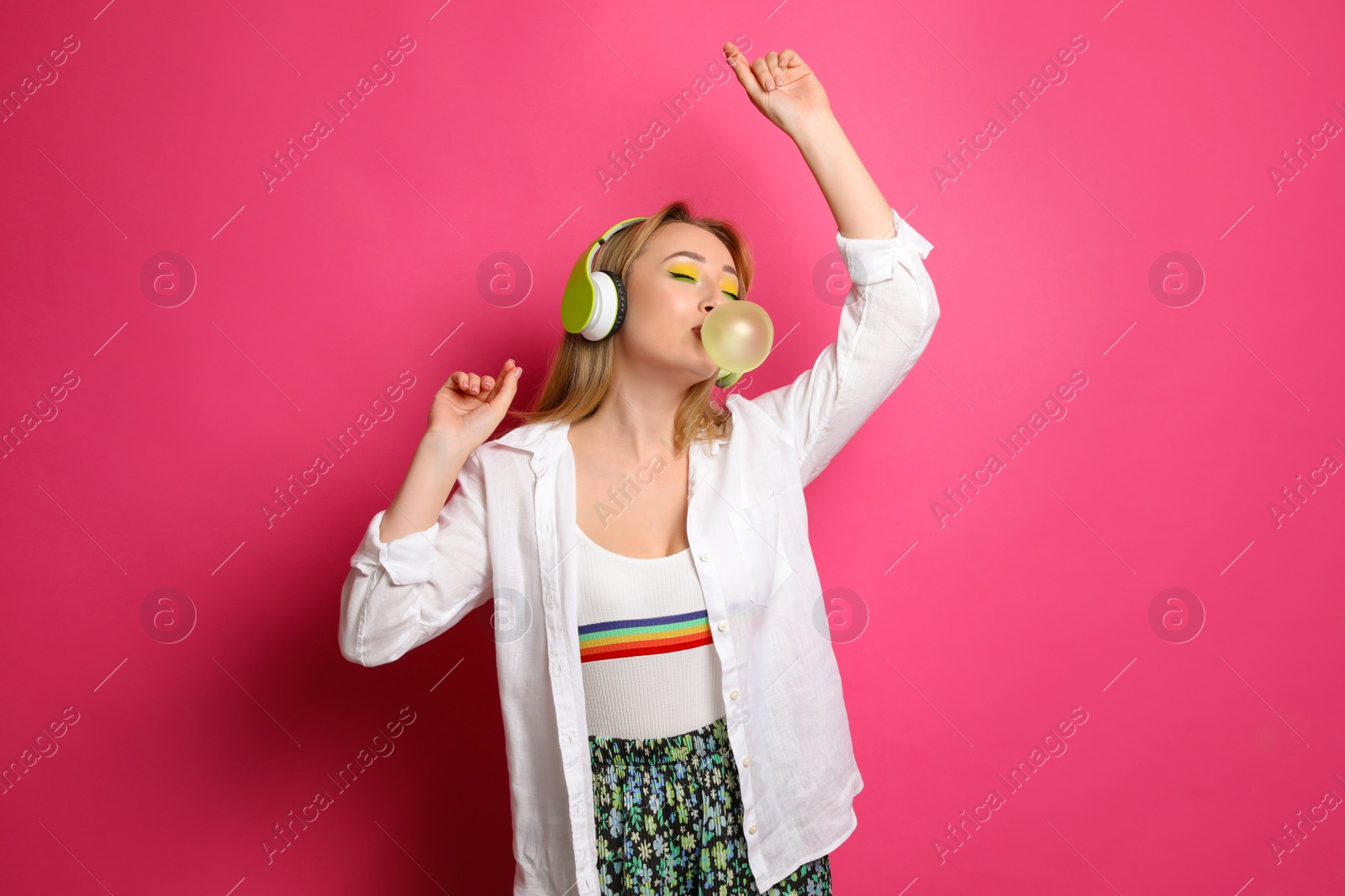 Photo of Fashionable young woman with bright makeup and headphones blowing bubblegum on pink background