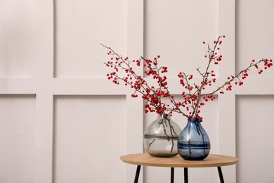 Photo of Hawthorn branches with red berries in vases on wooden table near light wall indoors, space for text