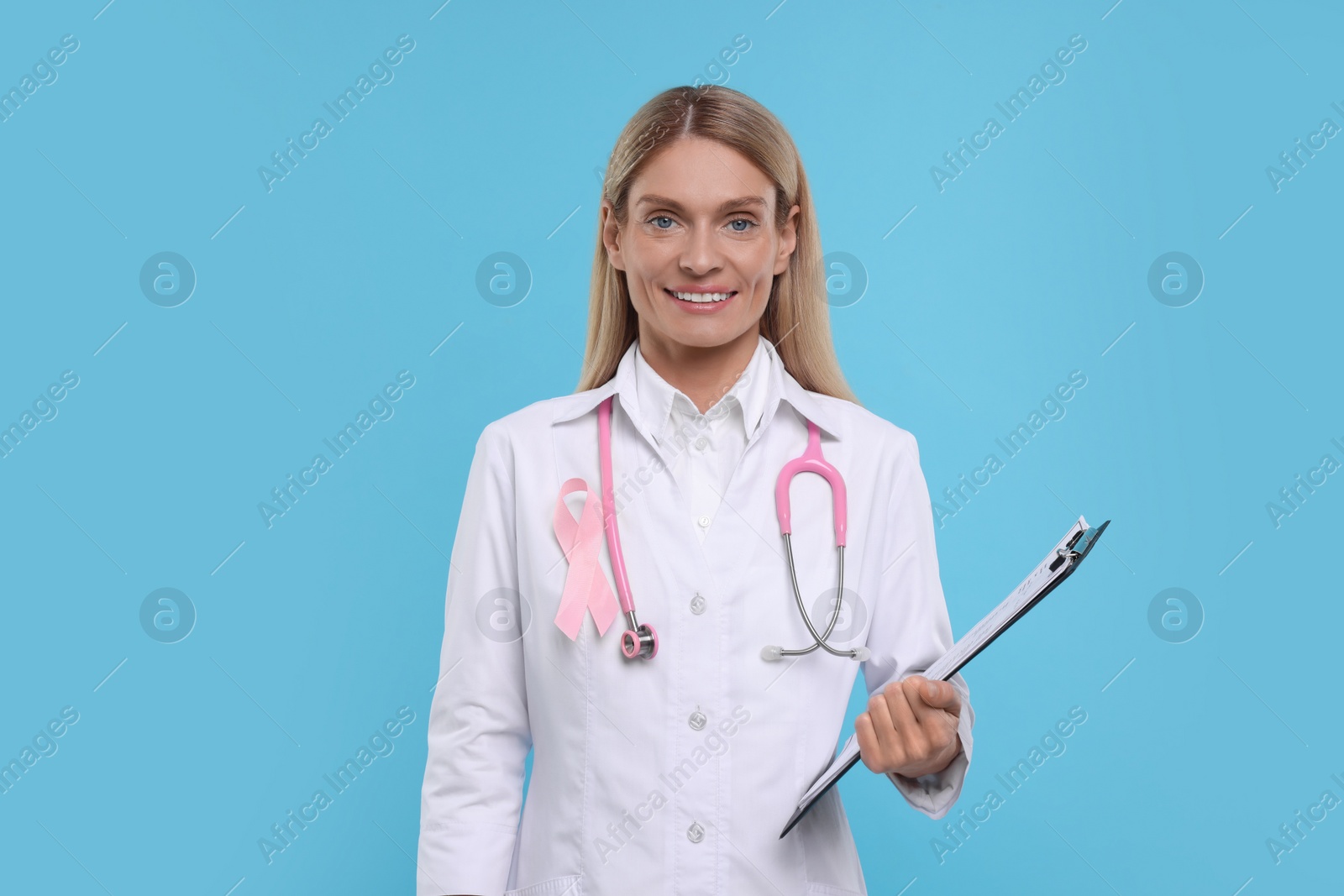 Photo of Doctor with pink ribbon, clipboard and stethoscope on light blue background. Breast cancer awareness