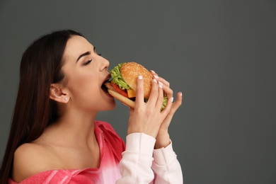 Young woman eating tasty burger on grey background. Space for text