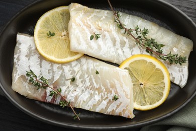 Photo of Fresh raw cod fillets with thyme and lemon in baking dish on table, top view
