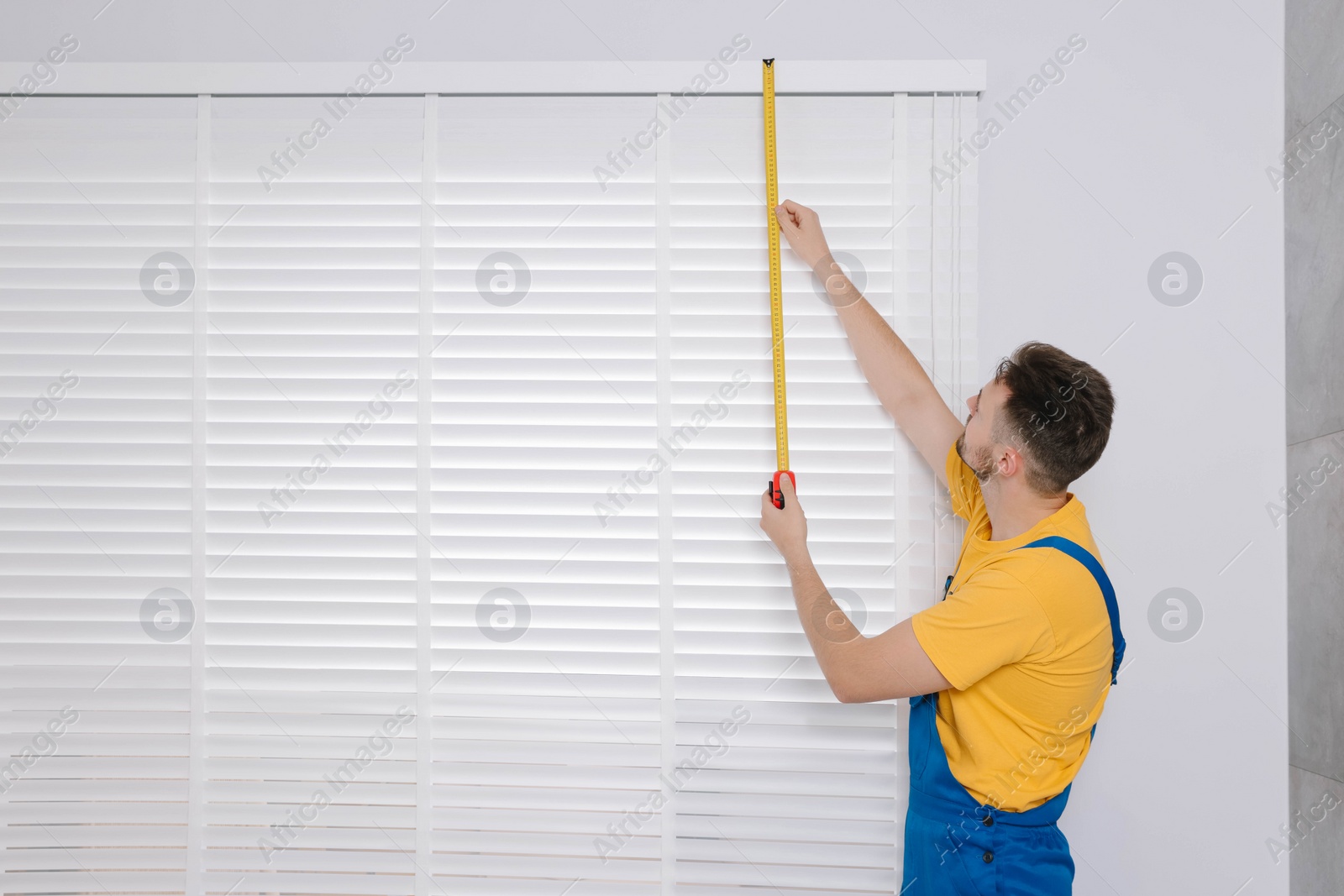 Photo of Worker in uniform using measuring tape while installing horizontal window blinds indoors