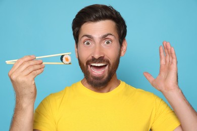 Photo of Emotional man holding sushi roll with chopsticks on light blue background