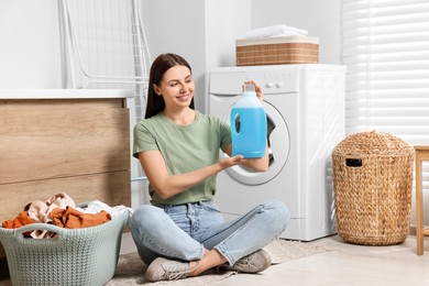 Woman sitting on floor near washing machine and holding fabric softener in bathroom