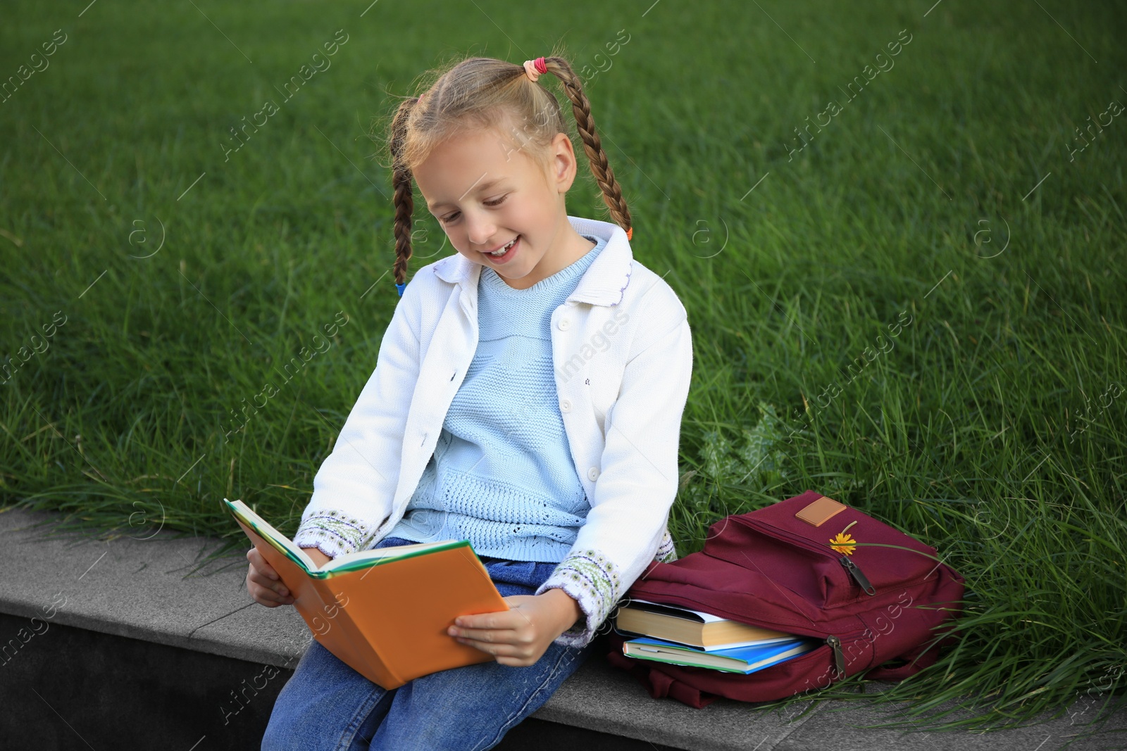 Photo of Cute little girl with backpack reading textbook on city street