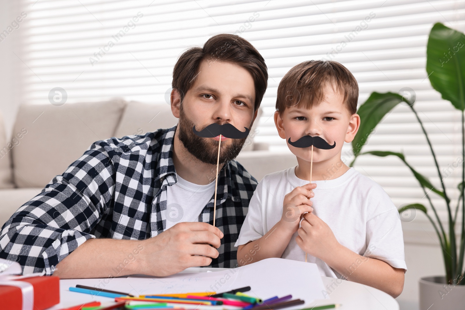 Photo of Dad and son covering mouth with paper mustaches at home