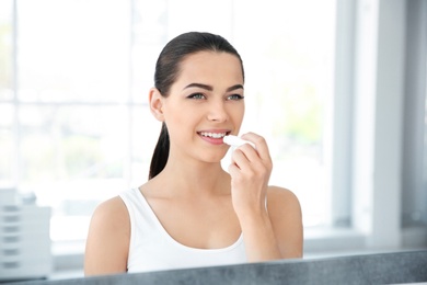 Photo of Young woman applying balm on her lips near mirror indoors