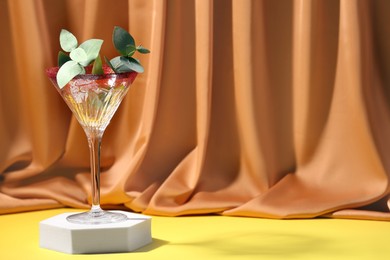 Beautiful martini glass with eucalyptus leaves on yellow surface near orange curtain. Space for text