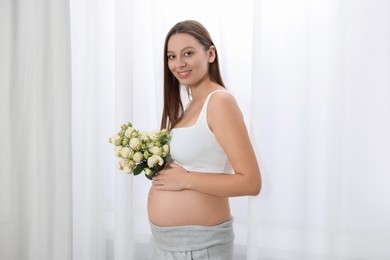 Beautiful pregnant woman with bouquet of roses near window indoors