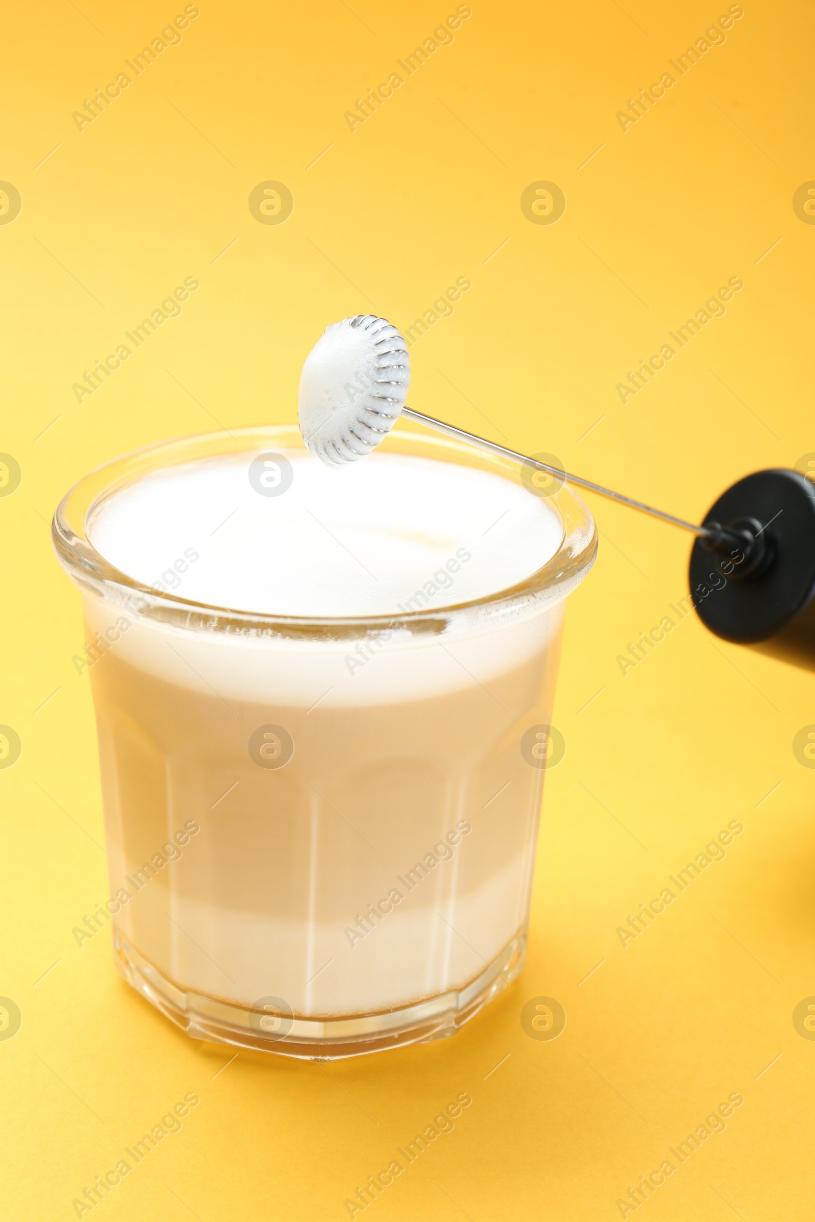 Photo of Mini mixer (milk frother) and tasty cappuccino in glass on yellow background