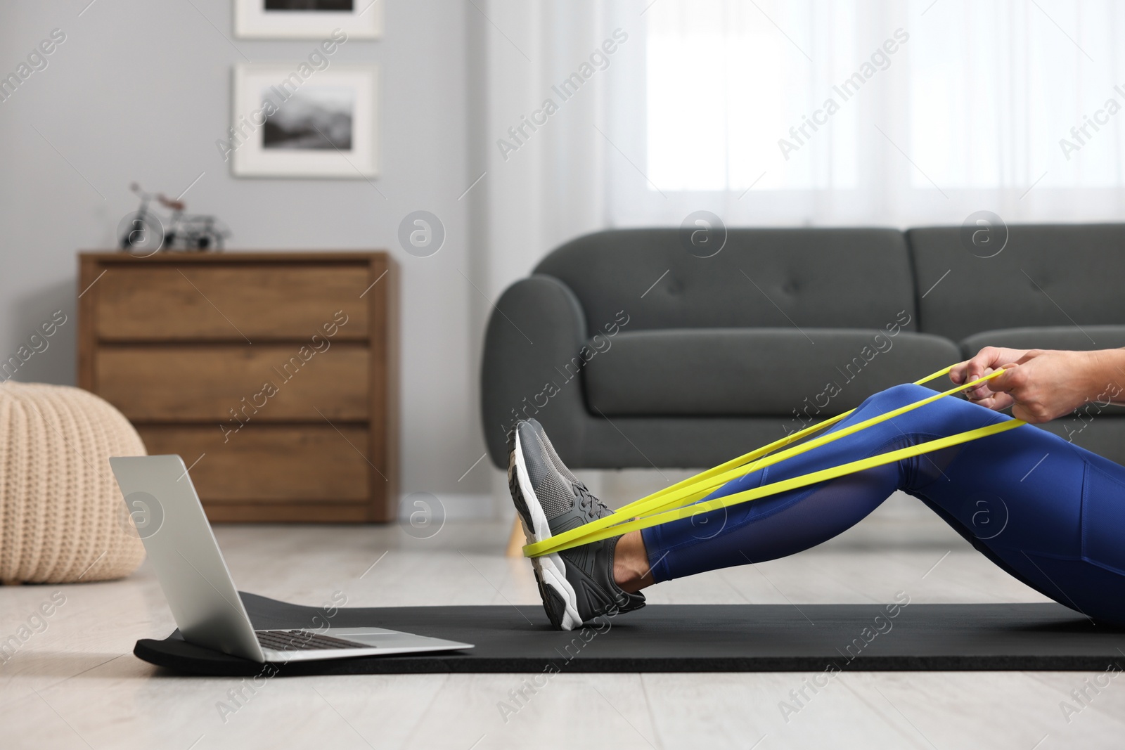 Photo of Woman doing exercise with fitness elastic band near laptop on mat at home, closeup