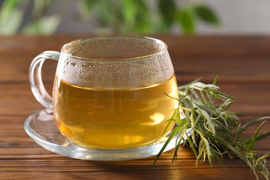 Photo of Cup of homemade herbal tea and fresh tarragon leaves on wooden table, closeup