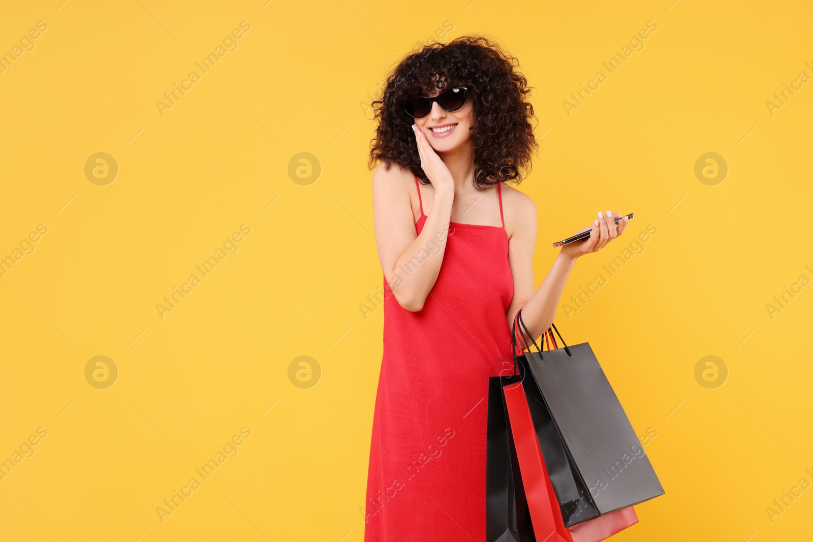 Photo of Happy young woman with shopping bags and modern smartphone on yellow background. Space for text