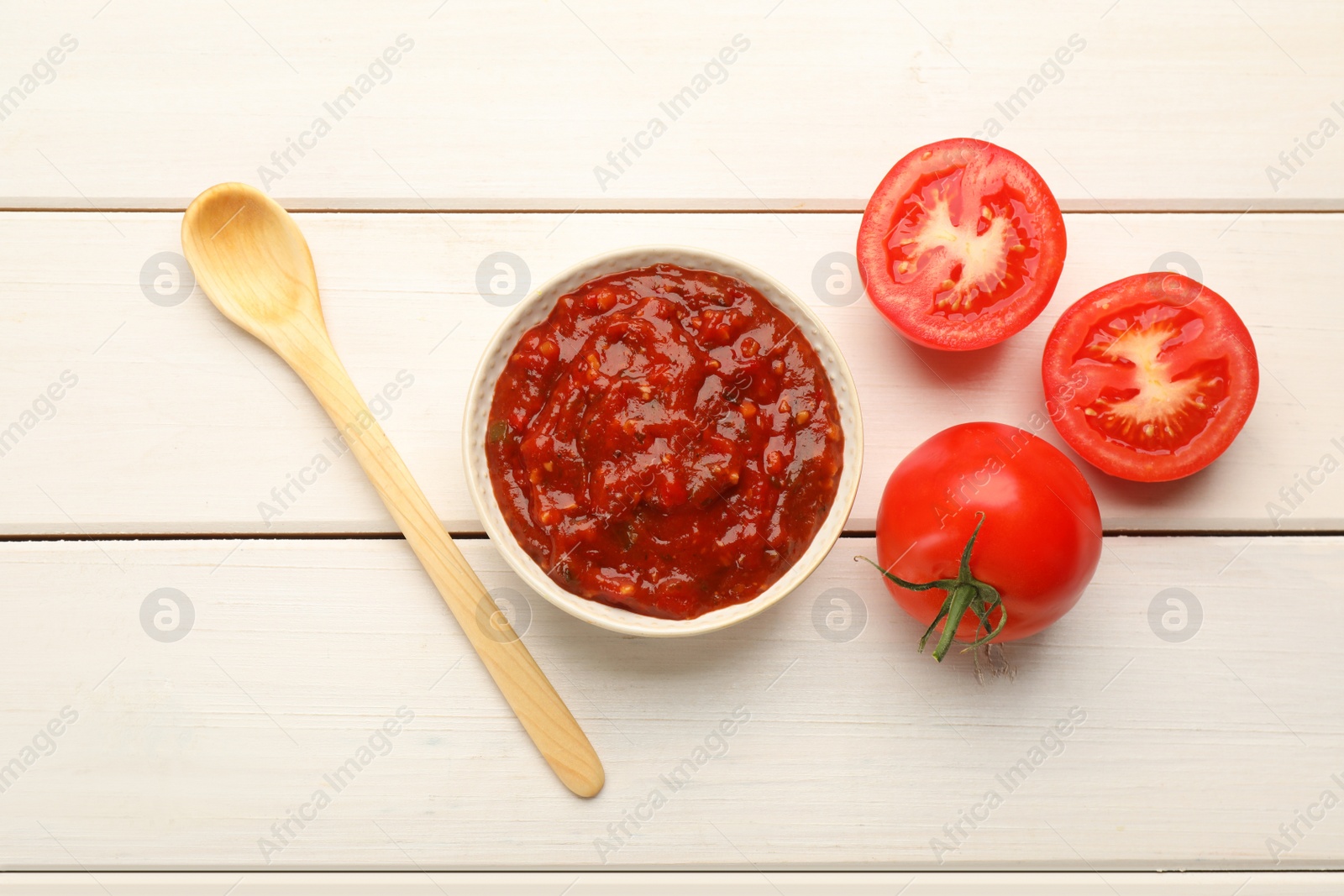Photo of Delicious adjika sauce in bowl with spoon and tomatoes on white wooden table, flat lay