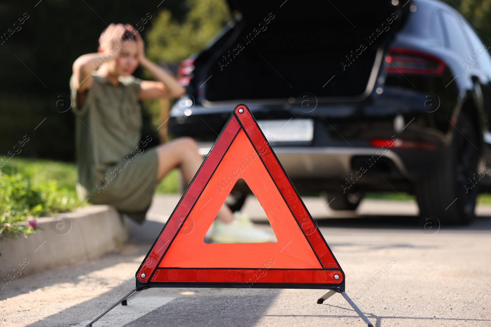 Photo of Woman sitting near broken car on roadside outdoors, focus on warning triangle