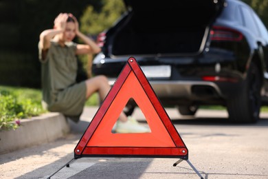 Photo of Woman sitting near broken car on roadside outdoors, focus on warning triangle