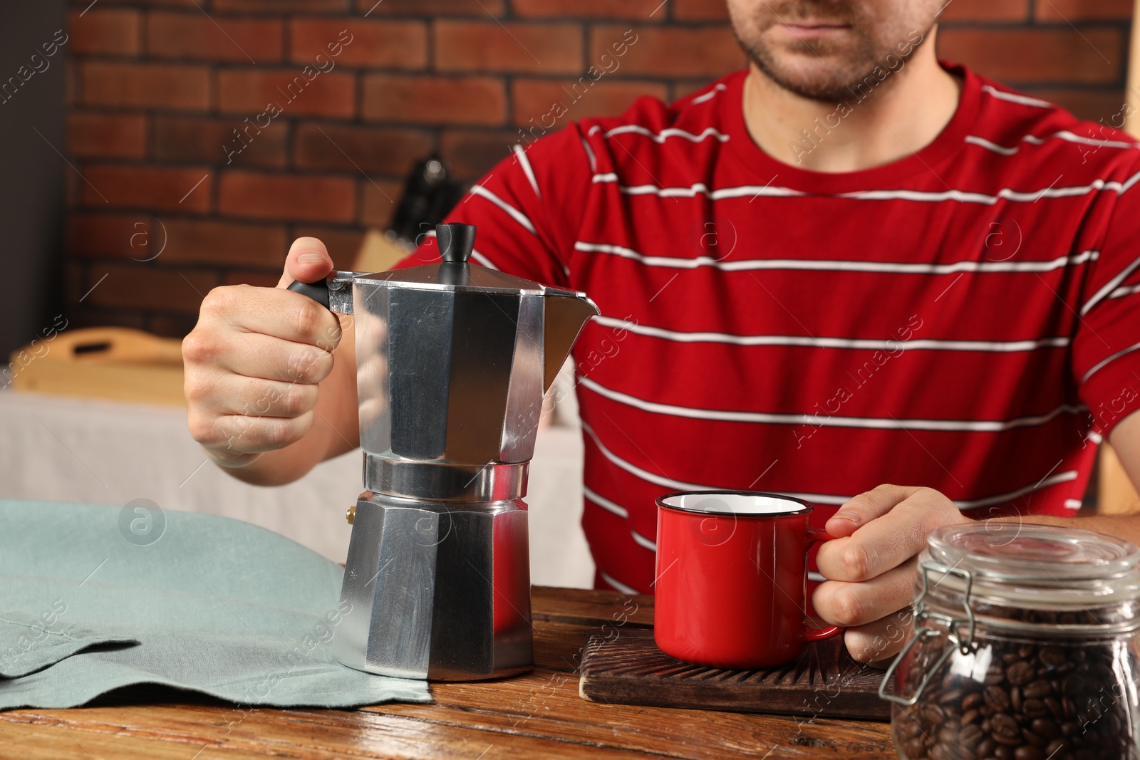 Photo of Brewing coffee. Man with jar of beans, moka pot and mug at wooden table indoors, closeup