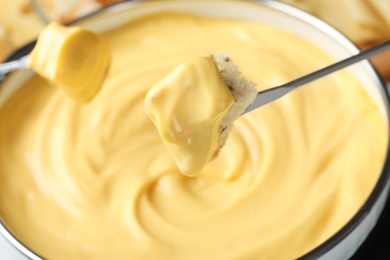 Photo of Piece of bread over pot with delicious cheese fondue, closeup