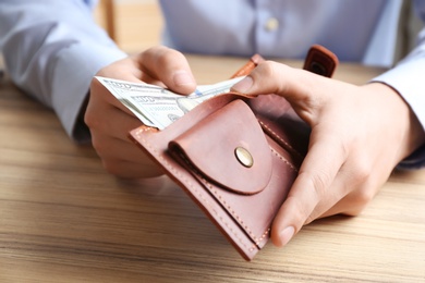 Photo of Man putting dollar banknotes into wallet at wooden table, closeup. Money savings