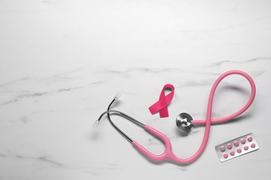 Breast cancer awareness. Pink ribbon, stethoscope and pills on white marble table, flat lay. Space for text