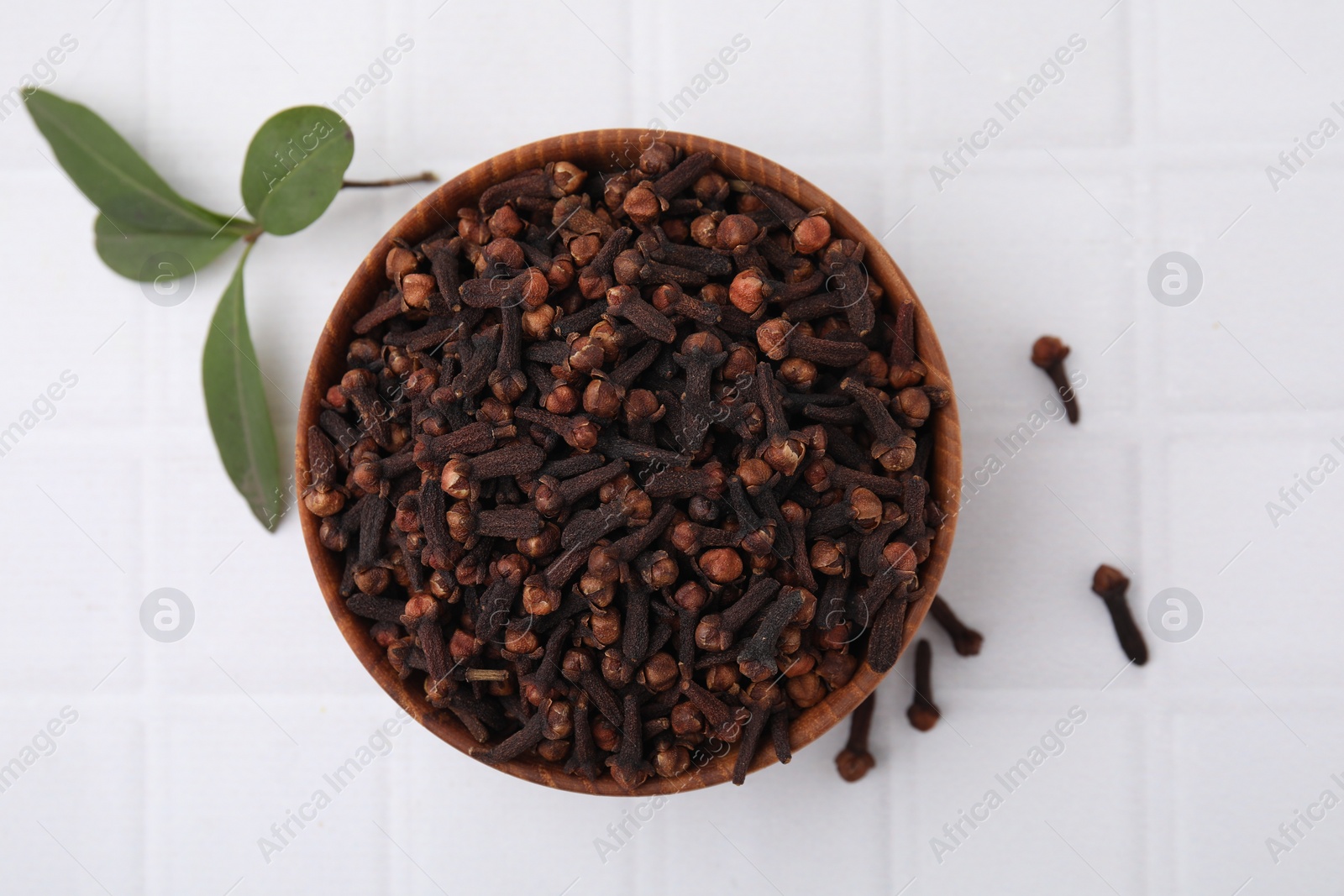 Photo of Aromatic cloves in bowl and green leaves on white tiled table, flat lay