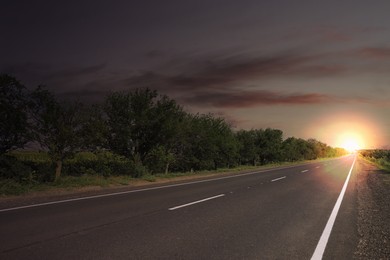 Empty asphalt road and trees under dark sky with rising sun