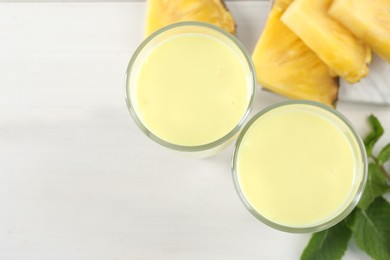 Tasty pineapple smoothie in glasses, mint and cut fruit on white wooden table, flat lay. Space for text