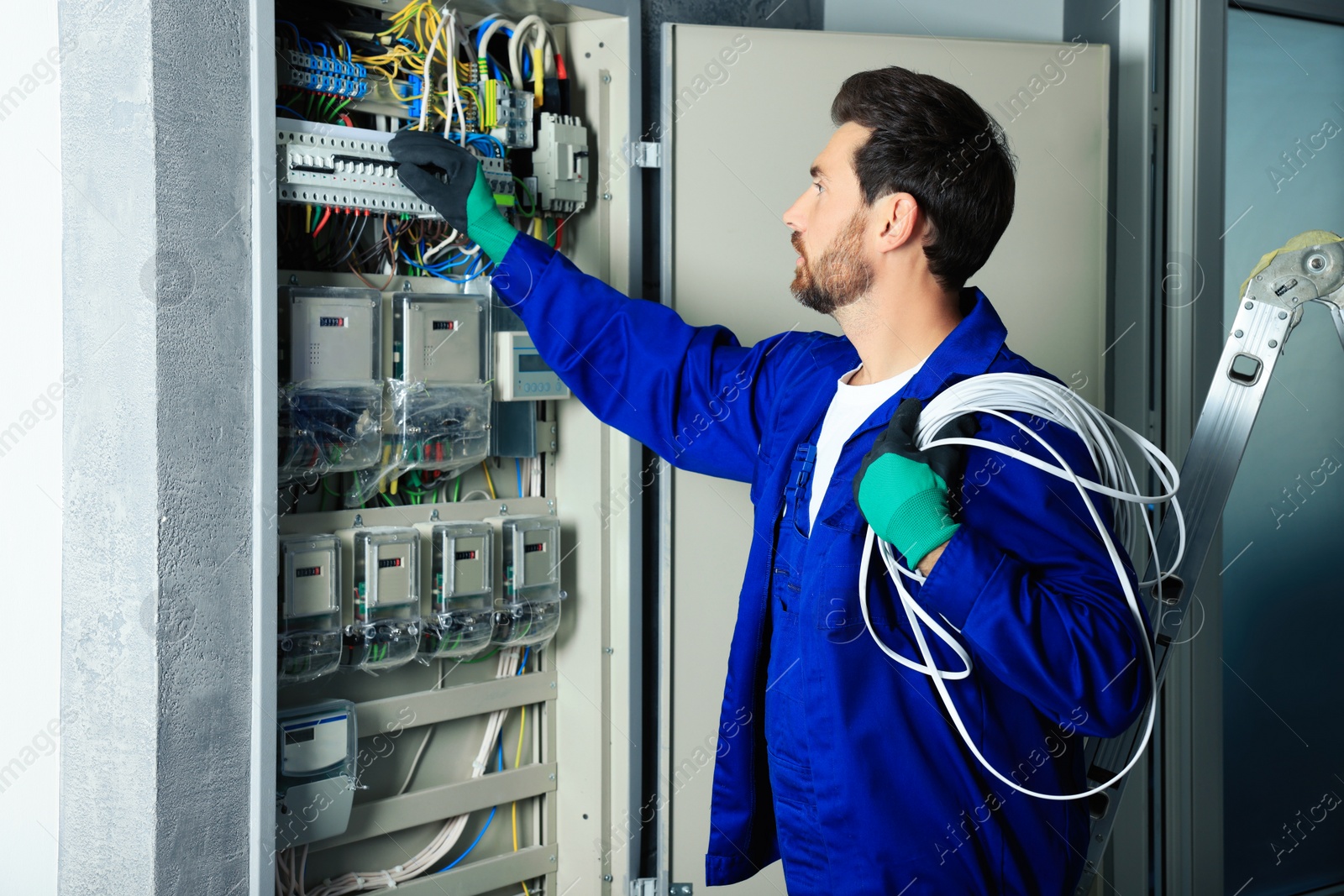 Photo of Electrician with wires switching off circuit breakers in fuse box indoors