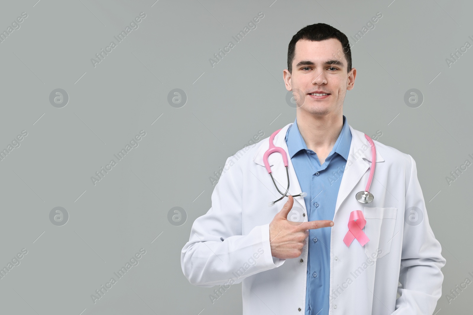 Photo of Portrait of smiling mammologist with stethoscope pointing at pink ribbon on grey background. Space for text