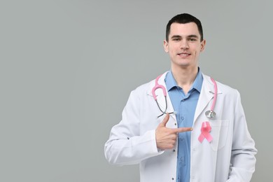 Portrait of smiling mammologist with stethoscope pointing at pink ribbon on grey background. Space for text