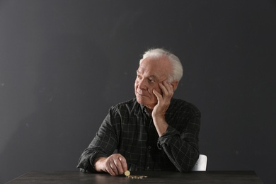 Poor elderly man with coins sitting at table on dark background