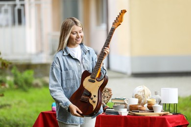 Photo of Woman holding guitar near table with different items on garage sale in yard