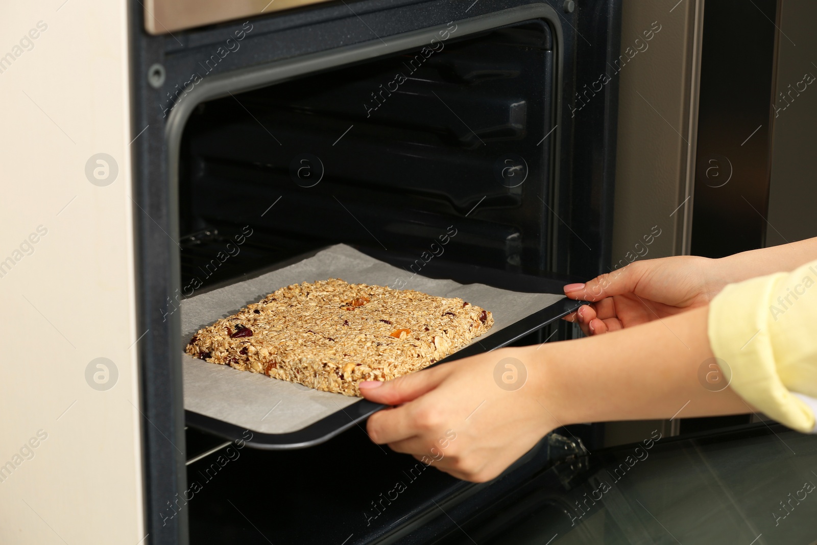 Photo of Making granola. Woman putting baking tray into oven in kitchen, closeup