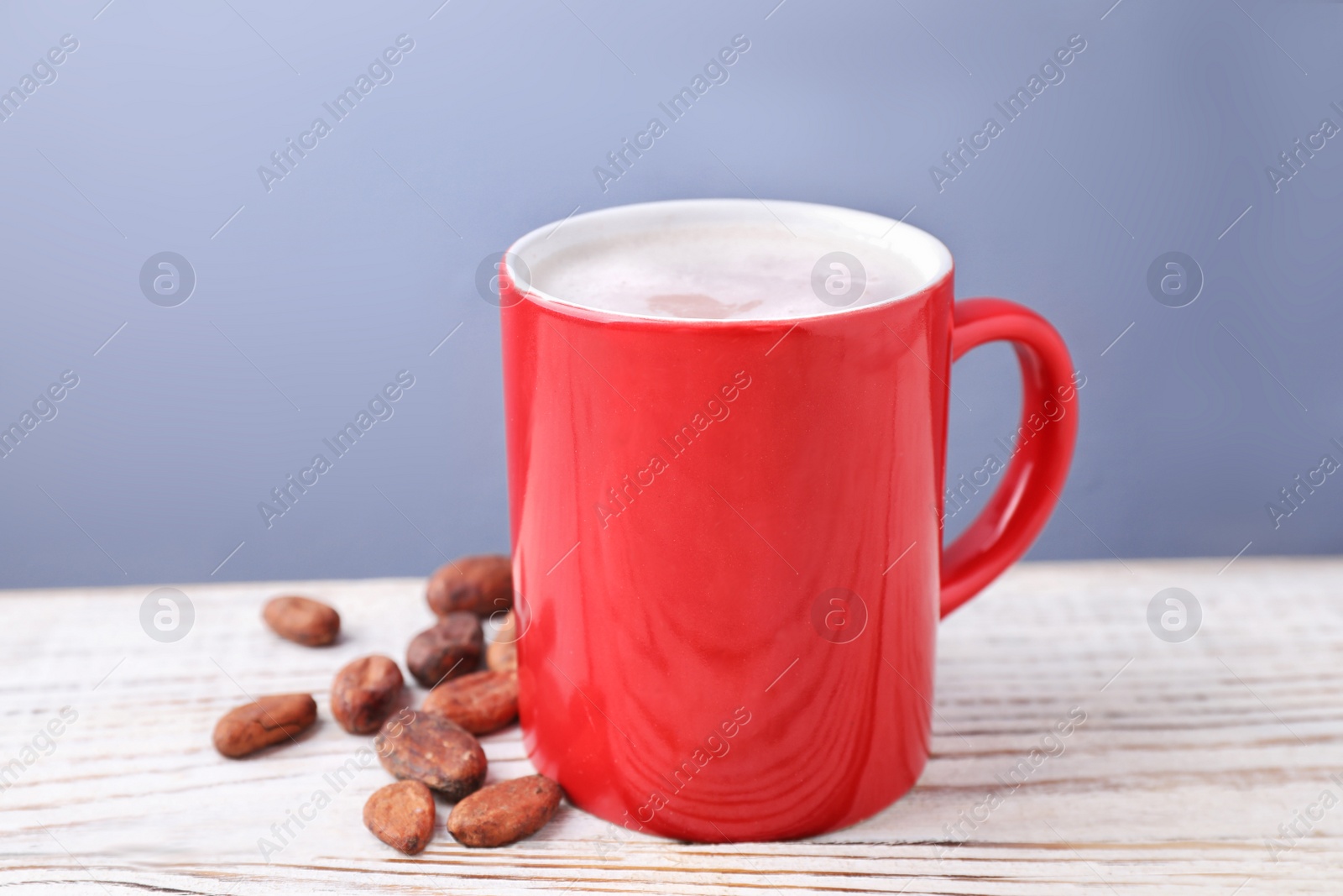 Photo of Cup of tasty cocoa and beans on white wooden table against grey background