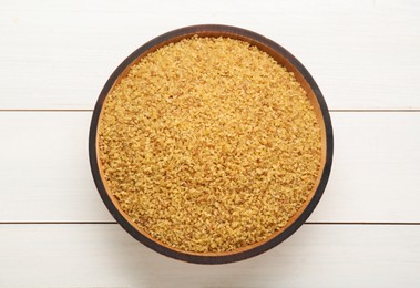 Bowl of uncooked bulgur on white wooden table, top view