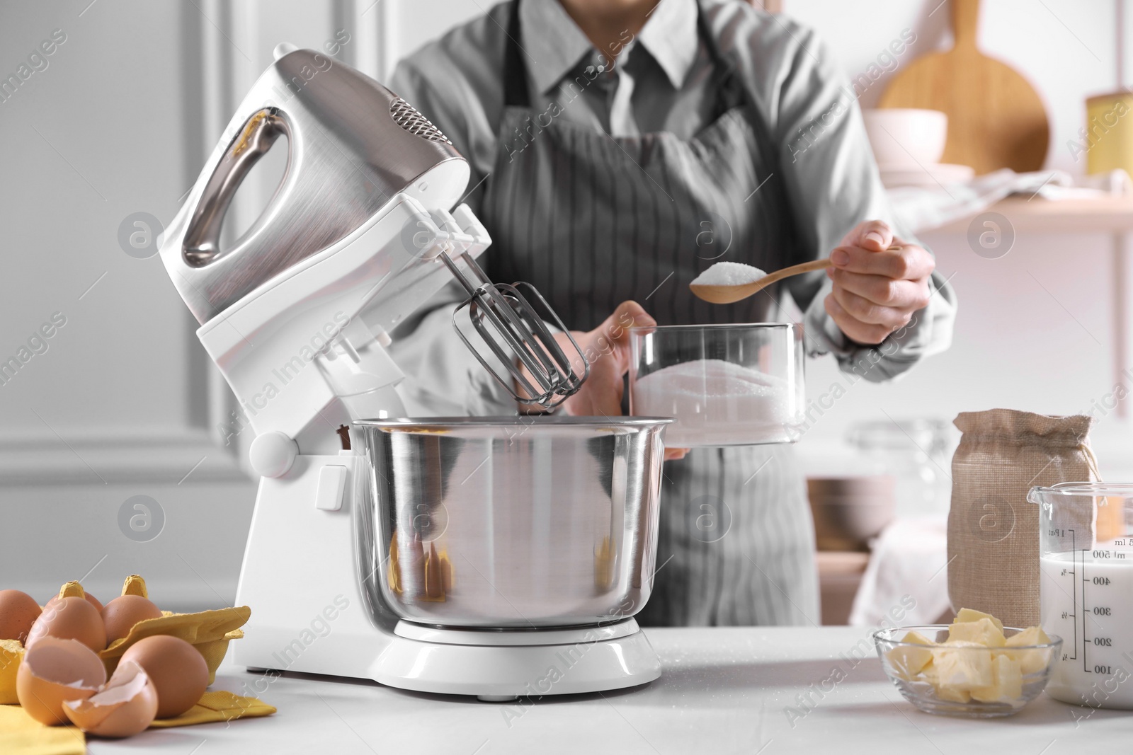 Photo of Woman adding sugar into bowl of stand mixer while making dough at table indoors, closeup