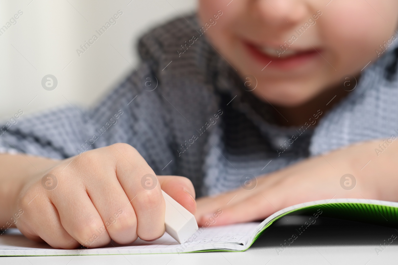 Photo of Little boy erasing mistake in his notebook at white desk, closeup