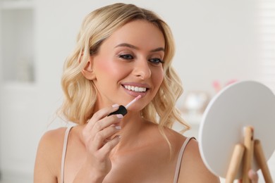 Photo of Beautiful makeup. Smiling woman with mirror applying liquid lipstick indoors