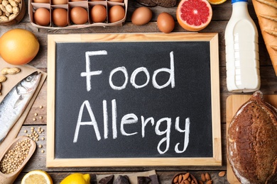 Photo of Chalkboard with words FOOD ALLERGY and different products on wooden table, flat lay