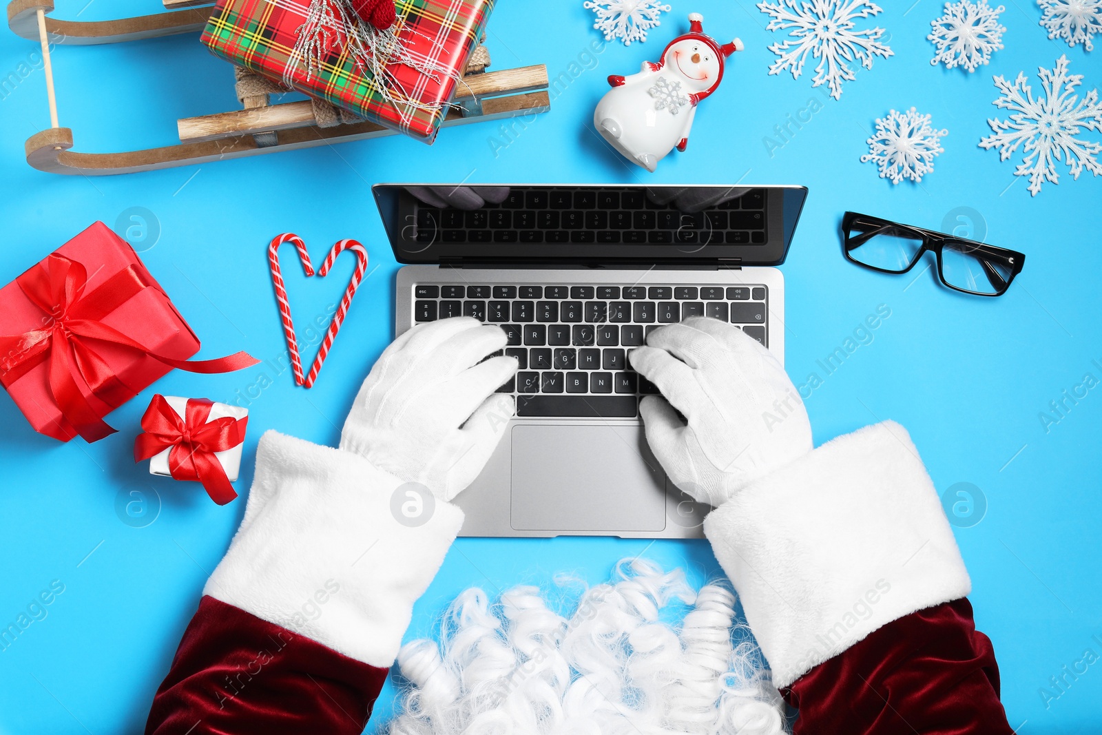 Photo of Santa Claus using laptop, closeup. Gift boxes, eyeglasses and Christmas decor on light blue background, top view